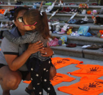 Emmy Morris who attended Indian day school and her mother who went to a residential school hugs her daughter Sharon Morris-Jones, 4, from the Tsartlip First Nation as they look at the Orange shirts, shoes, flowers and messages on display on the steps outside the legislature in Victoria, B.C., on June 8, 2021. Chad Hipolito/The Canadian Press.