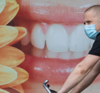 A man wearing a face mask to curb the spread of COVID-19 rides a bike past a photograph of a woman smiling outside a dental office, in Vancouver, B.C., Monday, Aug. 3, 2020. Darryl Dyck/The Canadian Press.