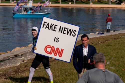 A man walks behind an on-air reporter holding a sign outside President Donald Trump's club, Mar-a-lago in Palm Beach, Tuesday, Nov. 15, 2022. Andrew Harnik/AP Photo.