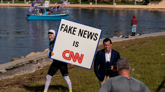 A man walks behind an on-air reporter holding a sign outside President Donald Trump's club, Mar-a-lago in Palm Beach, Tuesday, Nov. 15, 2022. Andrew Harnik/AP Photo.