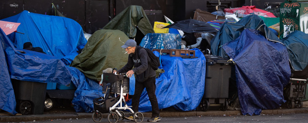 A man using a rolling walker walks on the street past tents setup on the sidewalk at a sprawling homeless encampment on East Hastings Street in the Downtown Eastside of Vancouver, on Tuesday, August 16, 2022. Darryl Dyck/The Canadian Press. 