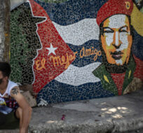 People rest in front of artwork by Cuban artist José Antonio Rodríguez Fuster featuring the late Fidel Castro, left, and Hugo Chavez, right, and the Spanish phrase "The best friend," in the seaside village of Jaimanitas on the outskirts of Havana, Cuba, Monday, May 10, 2021. Ramon Espinosa/AP Photo. 