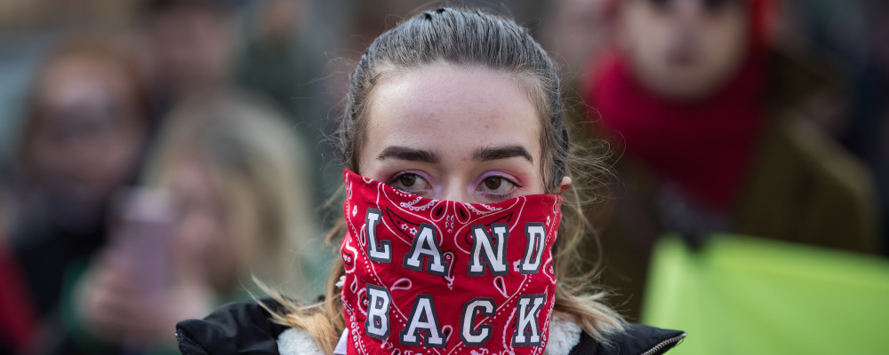 A woman wears a bandana over her face reading "Land Back" as protesters march to block a road used to access to the Port of Vancouver, during a demonstration in support of Wet'suwet'en Nation hereditary chiefs attempting to halt construction of a natural gas pipeline on their traditional territories, in Vancouver, on Monday, February 24, 2020. Darryl Dyck/The Canadian Press. 