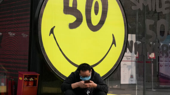 A man wearing a mask sits near a smiley face on Thursday, March 10, 2022, in Beijing. Ng Han Guan/AP Photo. 