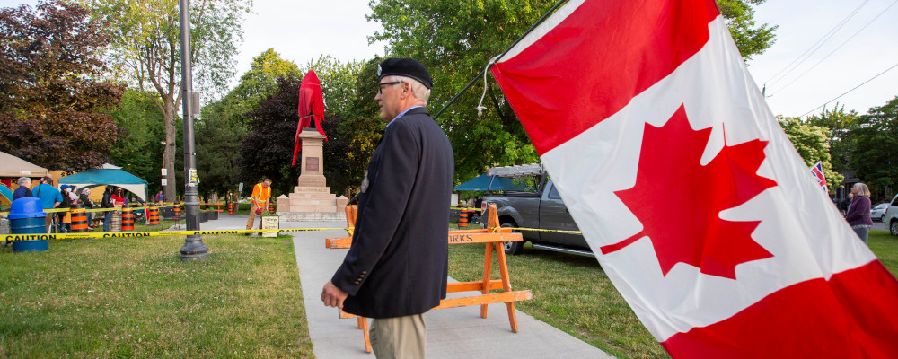 A person walks with a Canadian flag near the covered statue of Sir John A. Macdonald in Kingston, Ontario on Friday June 18, 2021.  Lars Hagberg/The Canadian Press. 